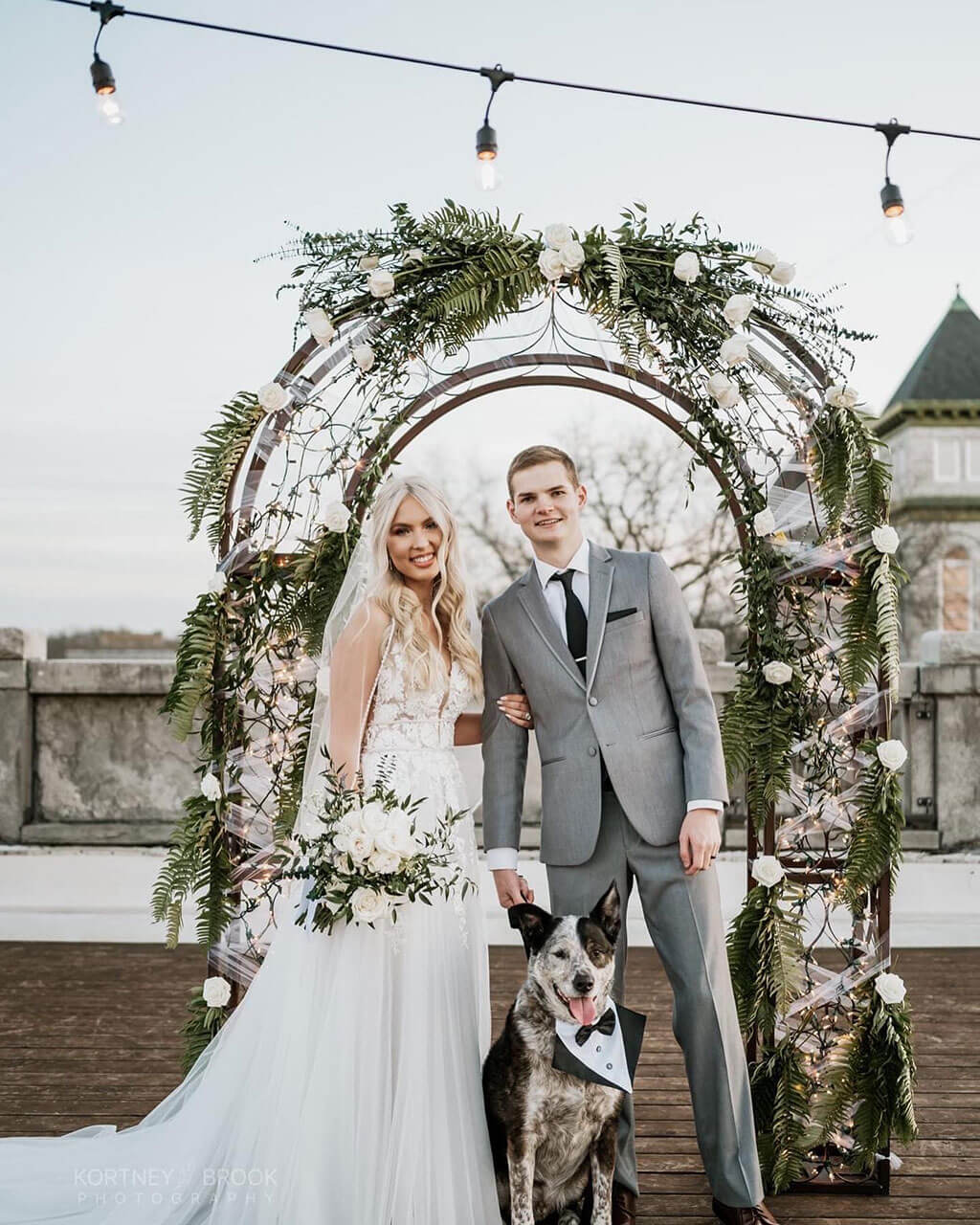 Bride and groom under a rose and fern arch on the rooftop with their dog