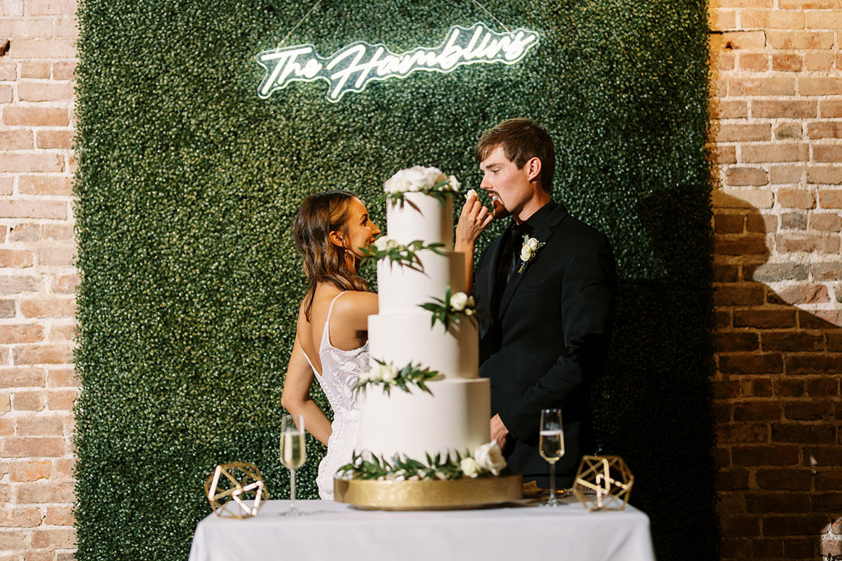 Couple cutting a tall tiered wedding cake in front of a greenery wall